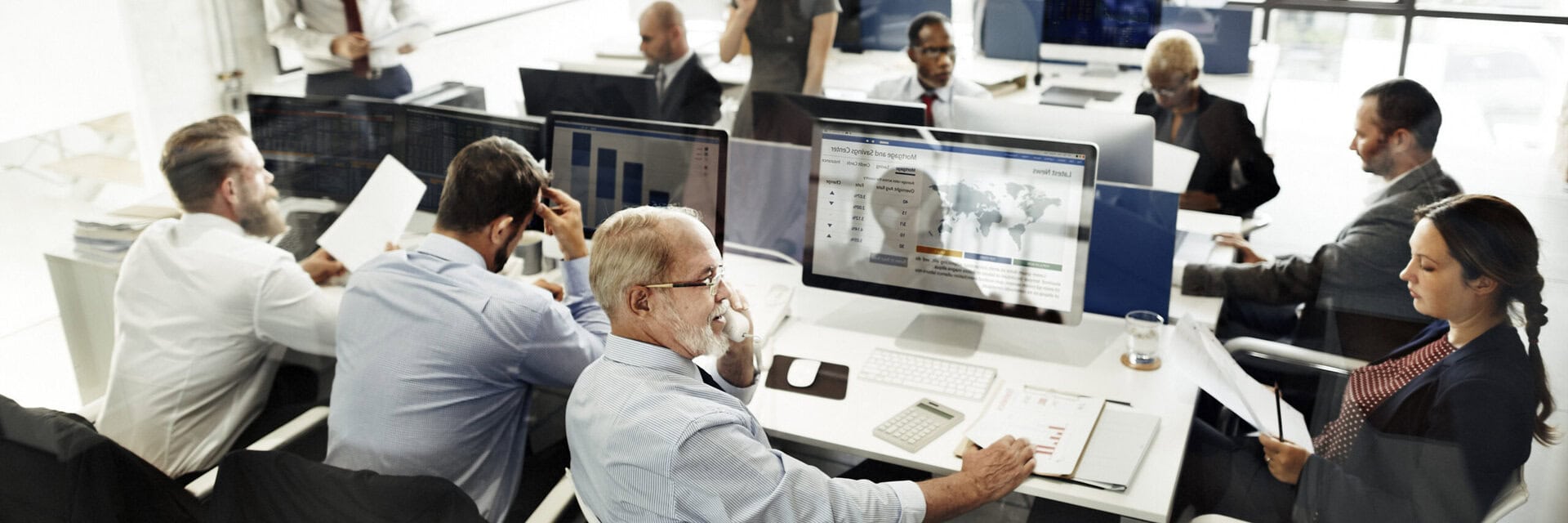 A group of people in business attire work together in a modern office, using computers, reviewing charts and documents, and having discussions around large desks with monitors and papers.
