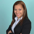 Woman with straight brown hair wearing a black blazer and striped shirt, standing against a light blue background, smiling with arms crossed.