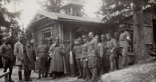 A group of soldiers in uniform stand outside a rustic wooden building surrounded by trees, posing for a photo during wartime.