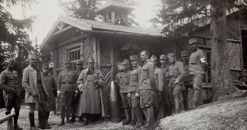 A group of soldiers in uniform stand outside a rustic wooden building surrounded by trees, posing for a photo during wartime.
