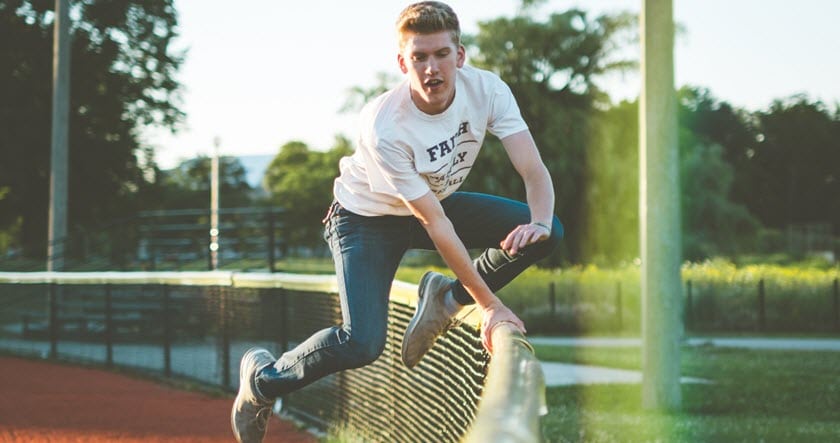 A young person in a white T-shirt and jeans jumps over a metal fence on an outdoor track during daylight.