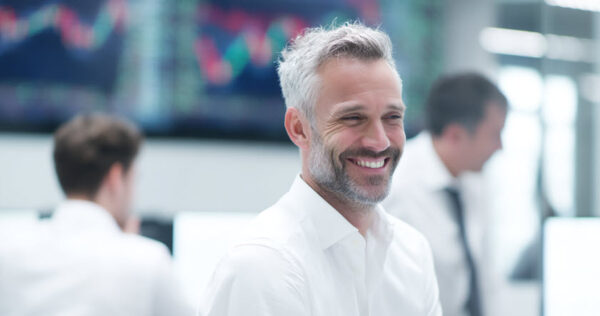 Smiling man with gray hair and beard in a white shirt stands in an office with blurred colleagues and computer screens in the background displaying graphs.