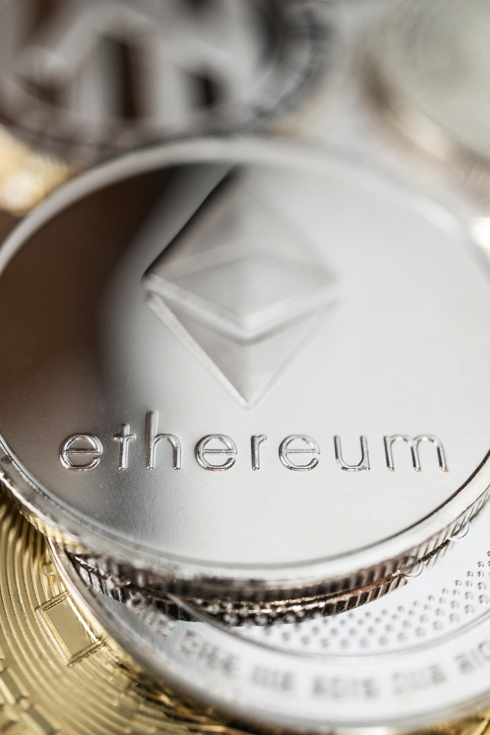 A close-up photo of a shiny silver Ethereum coin, displaying the Ethereum logo and the word ethereum engraved on its surface, with other blurred coins in the background.