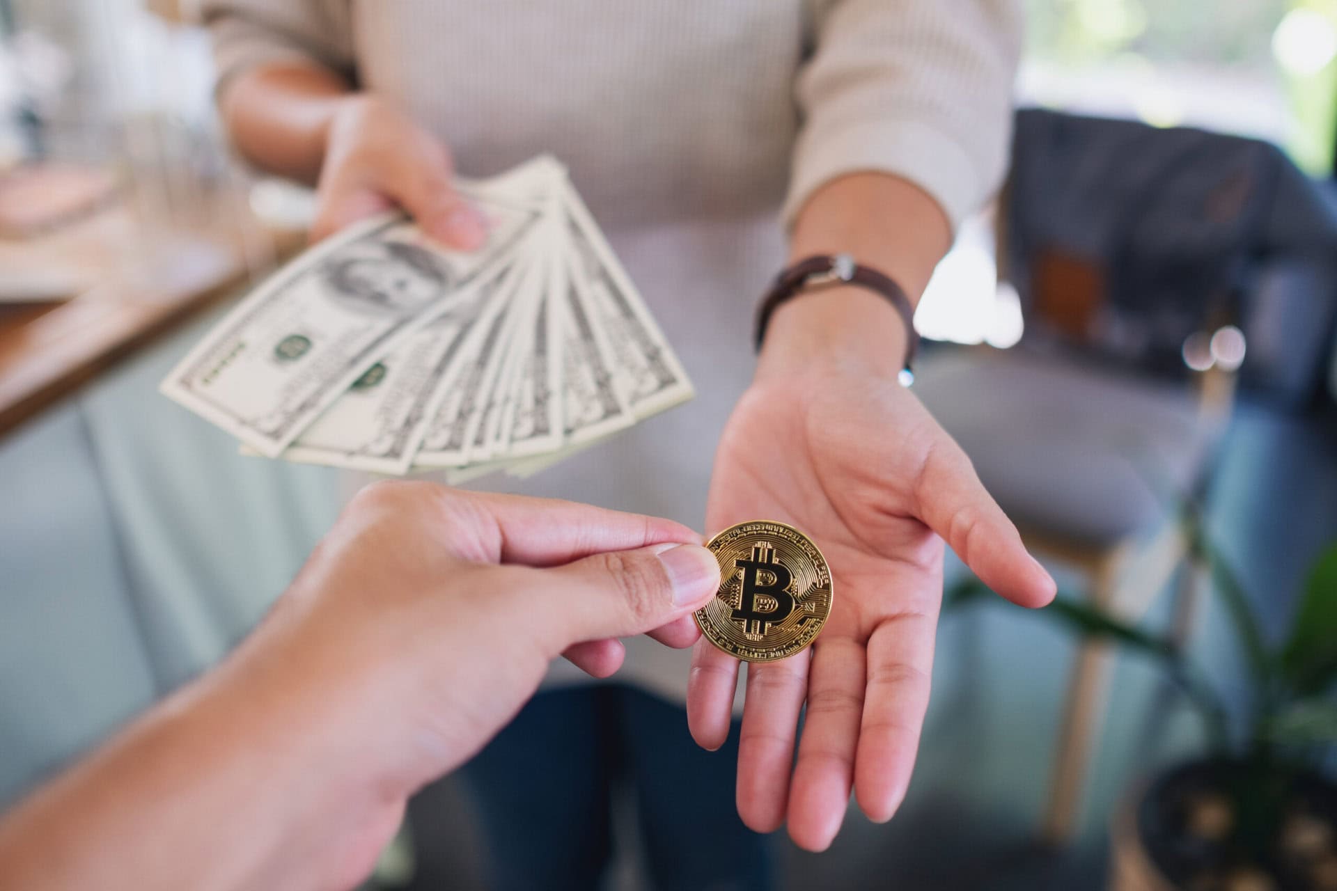 A person hands over several US dollar bills while another person gives them a gold Bitcoin coin, symbolizing an exchange between cryptocurrency and cash.