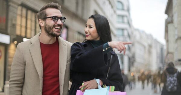A man and a woman walk together outdoors, smiling. The woman is holding shopping bags and pointing ahead while the man looks in the same direction.