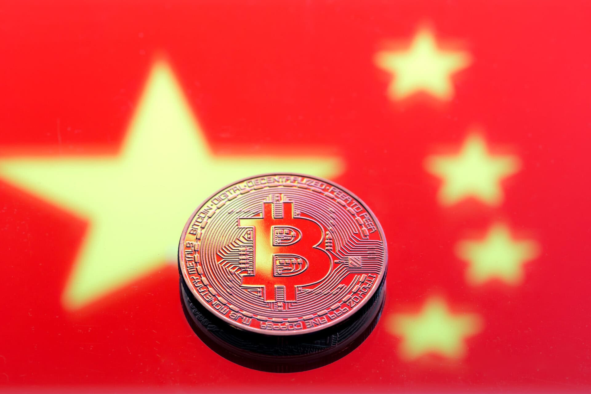 A physical Bitcoin token sits on a surface with the blurred Chinese flag in the background, featuring large and small yellow stars on a red field.