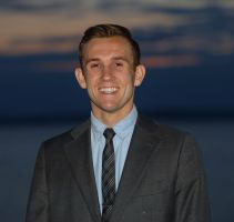 A young man in a suit and tie smiles at the camera with a calm body of water and a sunset sky in the background.