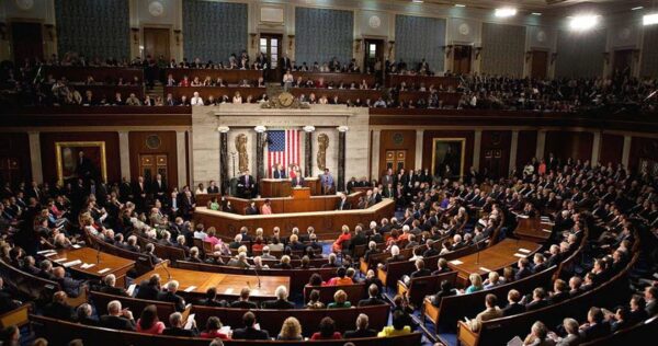 A large assembly in the U.S. House of Representatives chamber, with many people seated and an individual speaking at the podium in front of the American flag.