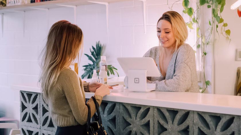 A woman stands at a counter interacting with a receptionist who is smiling and using a point-of-sale system in a brightly lit, modern space.