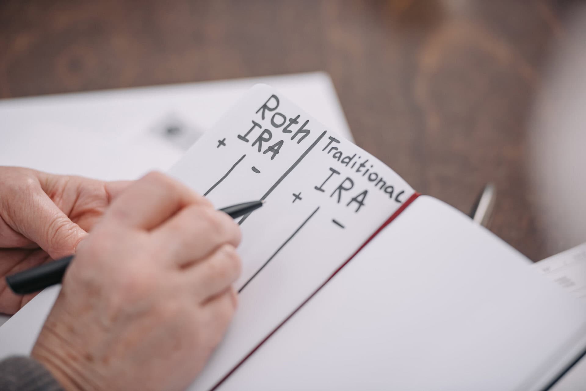 A person writes in a notebook, creating a comparison chart with columns labeled Roth IRA and Traditional IRA, each with plus and minus signs underneath.