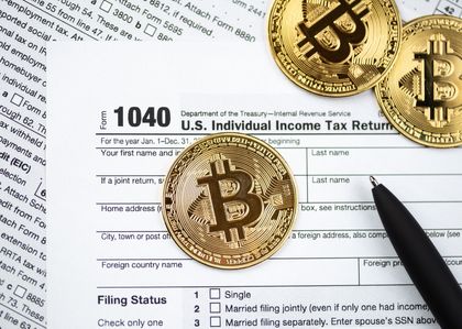 Three gold Bitcoin tokens rest on a U.S. 1040 income tax form, surrounded by tax documents, with a black pen nearby.
