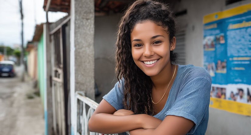A young woman with curly hair smiles while leaning on a fence outside, with colorful buildings and a blue poster in the background.