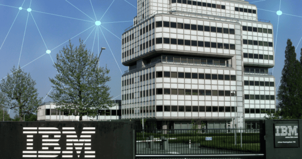 A modern IBM office building with a prominent IBM logo on a sign in the foreground; digital network graphics overlay the sky.