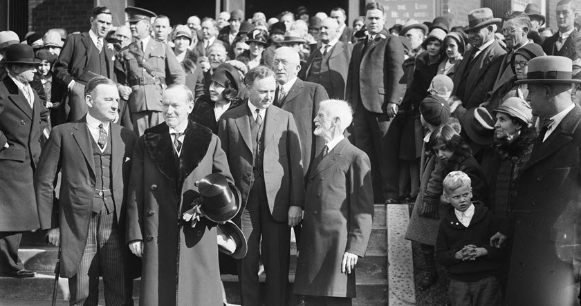 A large group of men and women, some in hats and overcoats, stand on exterior steps posing for a photo. Several men stand in front, with onlookers positioned on either side.