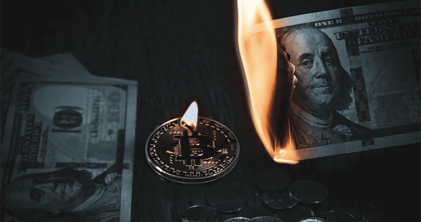A silver Bitcoin coin and stacks of coins are next to a burning US $100 bill, with more US bills partially visible in the background.