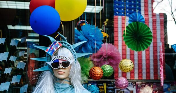 A mannequin dressed as the Statue of Liberty with sunglasses and balloons stands in front of a shop decorated with colorful paper ornaments and an American flag.