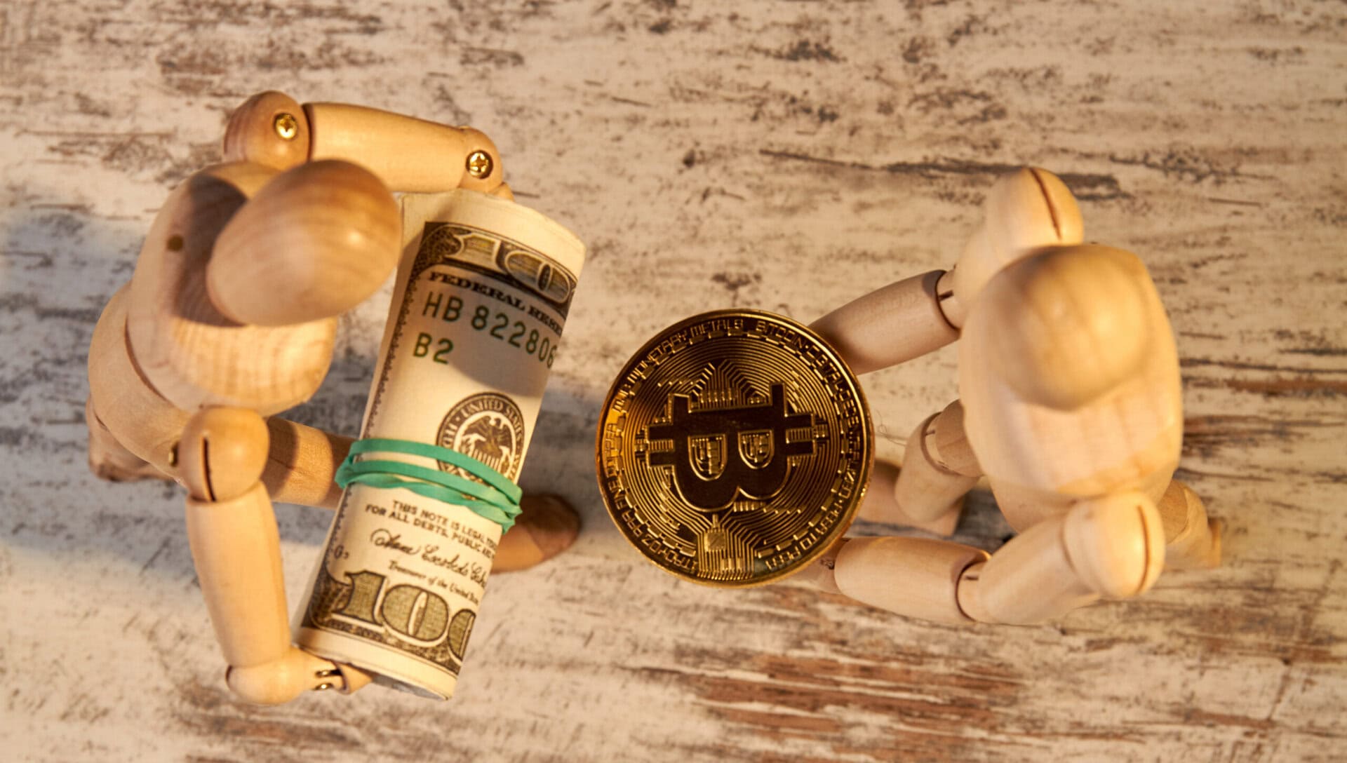Two wooden mannequins face each other; one holds a roll of US dollar bills while the other holds a golden Bitcoin, symbolizing the exchange between traditional currency and cryptocurrency.