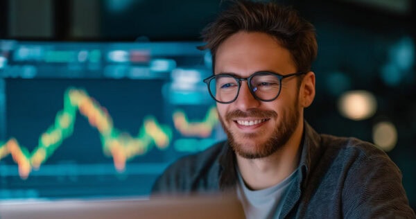 A man with glasses smiles while looking at a laptop, with a large financial chart displaying fluctuating data in the background.
