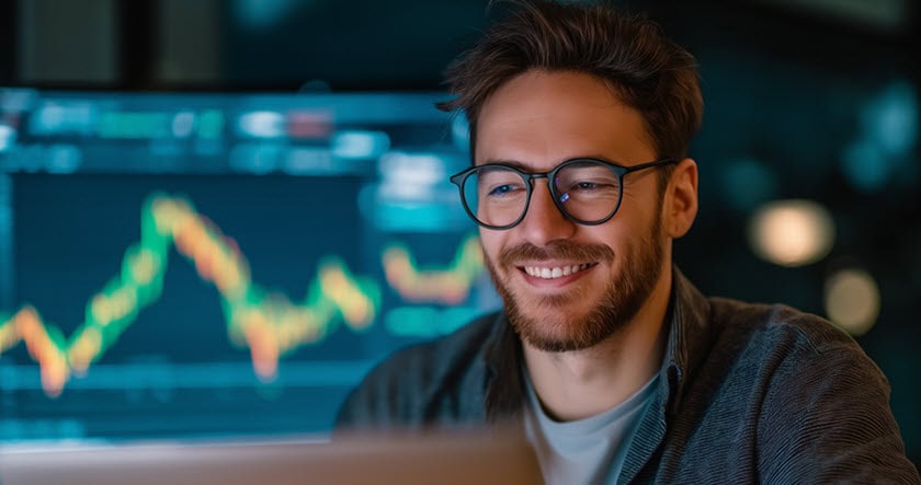 A man with glasses smiles while looking at a laptop, with a large financial chart displaying fluctuating data in the background.