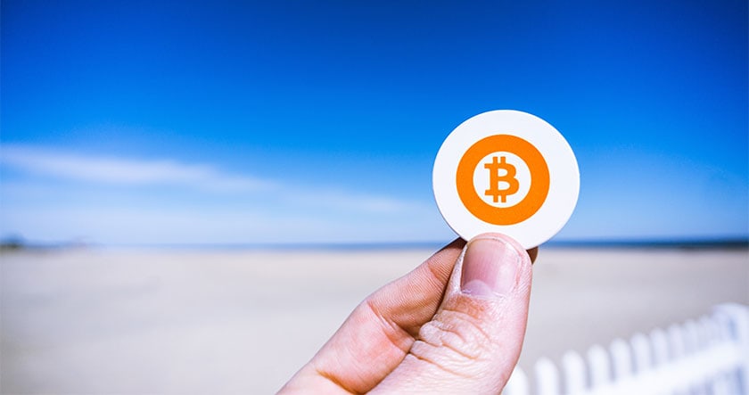 A hand holds a round token with a Bitcoin symbol against a blurred background of a beach and blue sky.
