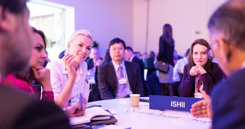 A group of professionals sit around a table in discussion at a conference, with a nameplate labeled "ISHII" visible on the table.