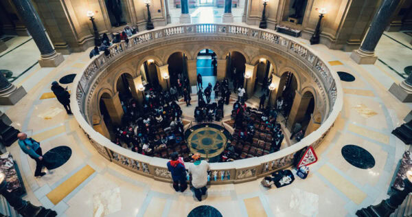 People gathered inside a grand, circular rotunda with marble floors and ornate columns, viewed from above.