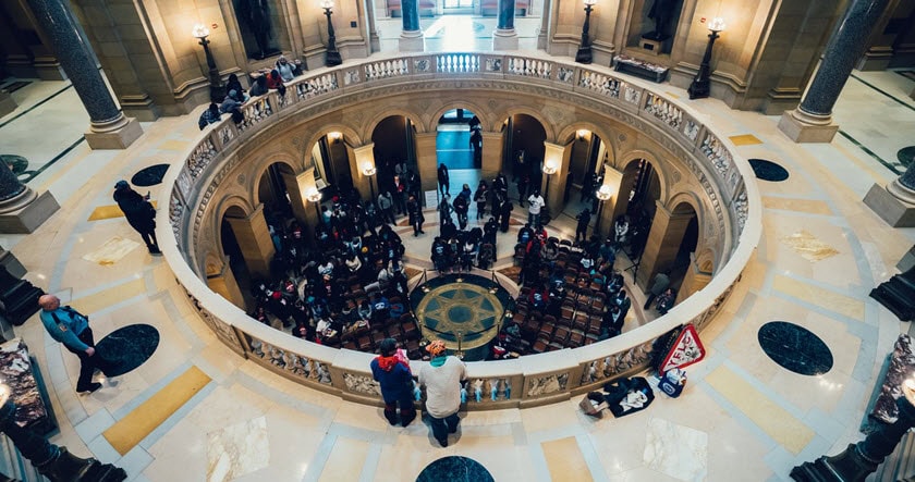 People gathered inside a grand, circular rotunda with marble floors and ornate columns, viewed from above.