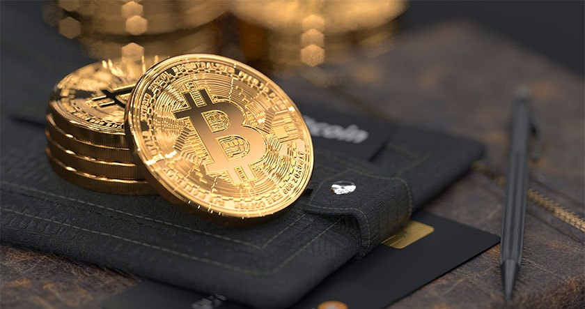 A stack of gold-colored physical Bitcoin tokens placed on a leather wallet, with credit cards and a pen nearby.