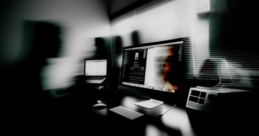 A dimly lit workspace with two computer screens on a desk; one monitor displays a blurry face, and shadowy figures appear in the background.
