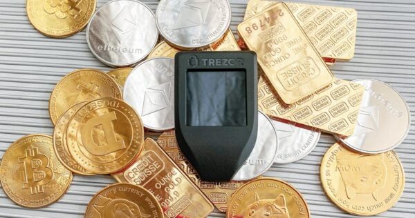A Trezor hardware wallet sits on a pile of gold and silver cryptocurrency coins and gold bars, including Bitcoin, Dogecoin, and Ethereum tokens.