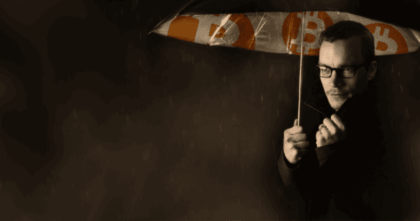 A man wearing glasses holds an umbrella with Bitcoin symbols, sheltering from rain in a dimly lit setting.