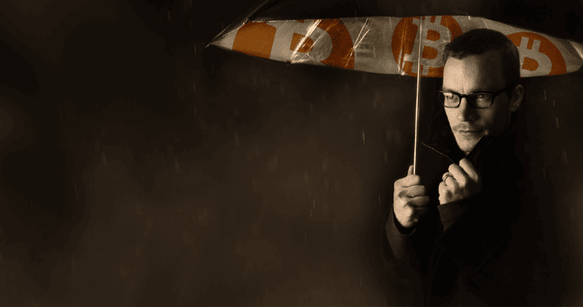 A man wearing glasses holds an umbrella with Bitcoin symbols, sheltering from rain in a dimly lit setting.
