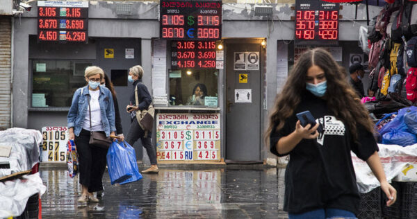 People walk past a currency exchange booth displaying exchange rates in a busy outdoor market area; some are wearing masks and carrying shopping bags.