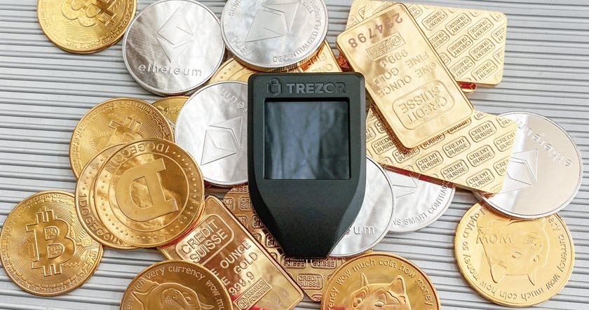 A Trezor hardware wallet sits on a pile of gold and silver cryptocurrency coins and gold bars, including Bitcoin, Dogecoin, and Ethereum tokens.