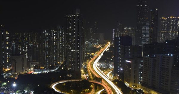 Cityscape at night showing tall buildings and illuminated highways with light trails from moving vehicles.