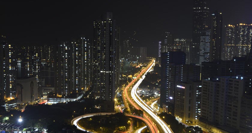 Cityscape at night showing tall buildings and illuminated highways with light trails from moving vehicles.