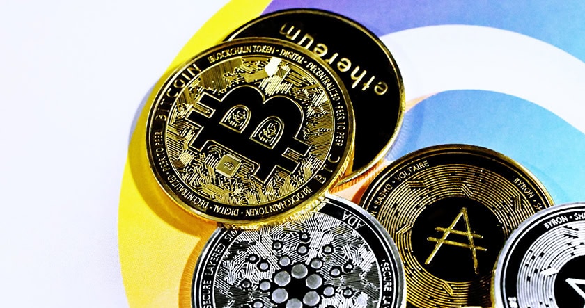 Several physical coins representing different cryptocurrencies, including Bitcoin, Ethereum, Cardano, and Algorand, are arranged on a colorful background.