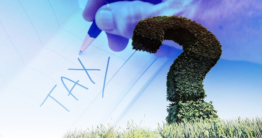 A large question mark-shaped hedge stands in grass; overlaid is a hand writing "TAX" on paper, suggesting tax-related uncertainty or questions.