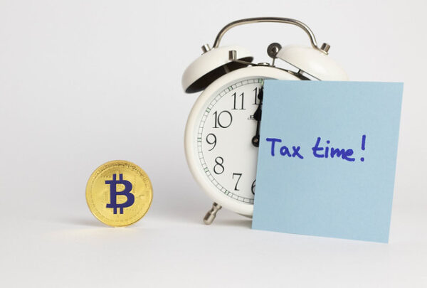 A Bitcoin token, an alarm clock, and a note with “Tax time!” written on it are arranged on a white surface.