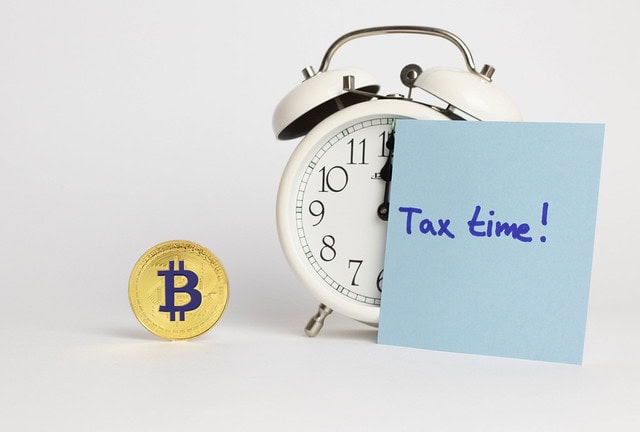 A Bitcoin token, an alarm clock, and a note with “Tax time!” written on it are arranged on a white surface.