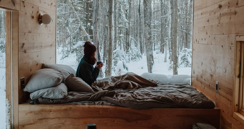 A person sits on an unmade bed next to a large window, looking out at a snowy forest. The room has wooden walls and a cozy atmosphere.