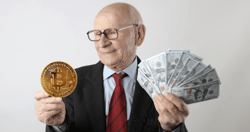 An older man in a suit holds a large mock Bitcoin coin in one hand and several U.S. hundred-dollar bills in the other, standing against a plain background.