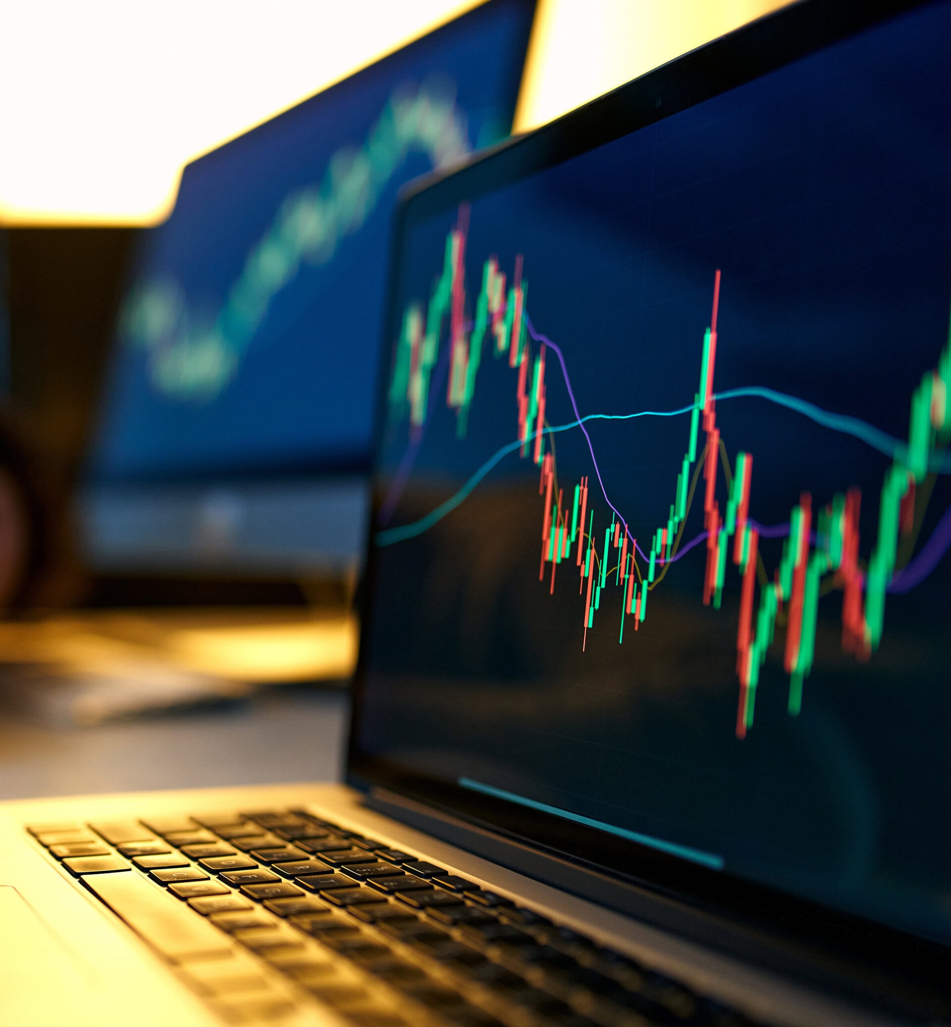 A close-up of a laptop screen displaying colorful candlestick charts and trend lines, indicating financial market data analysis, with another blurred screen showing a similar chart in the background.