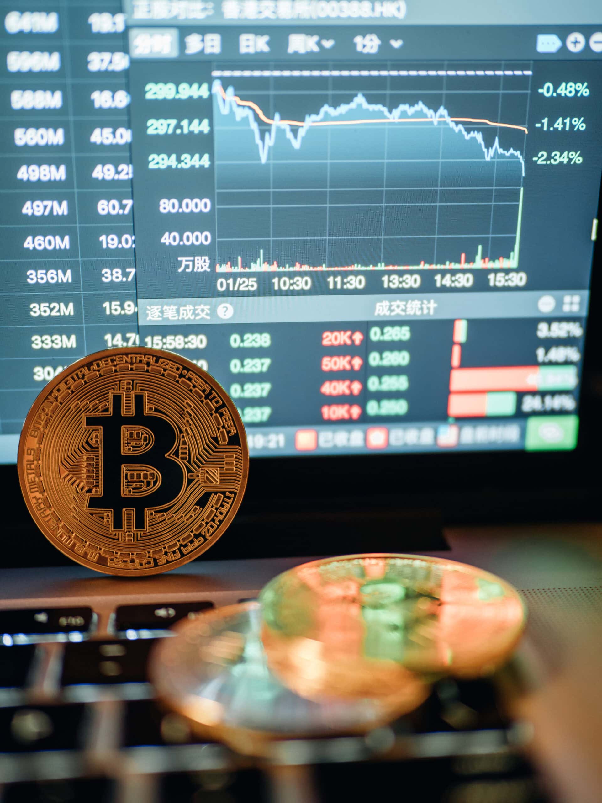 A physical Bitcoin coin sits on a laptop keyboard with a financial chart and stock data displayed on the screen in the background. The chart shows a downward trend, and text on the monitor is in Chinese.