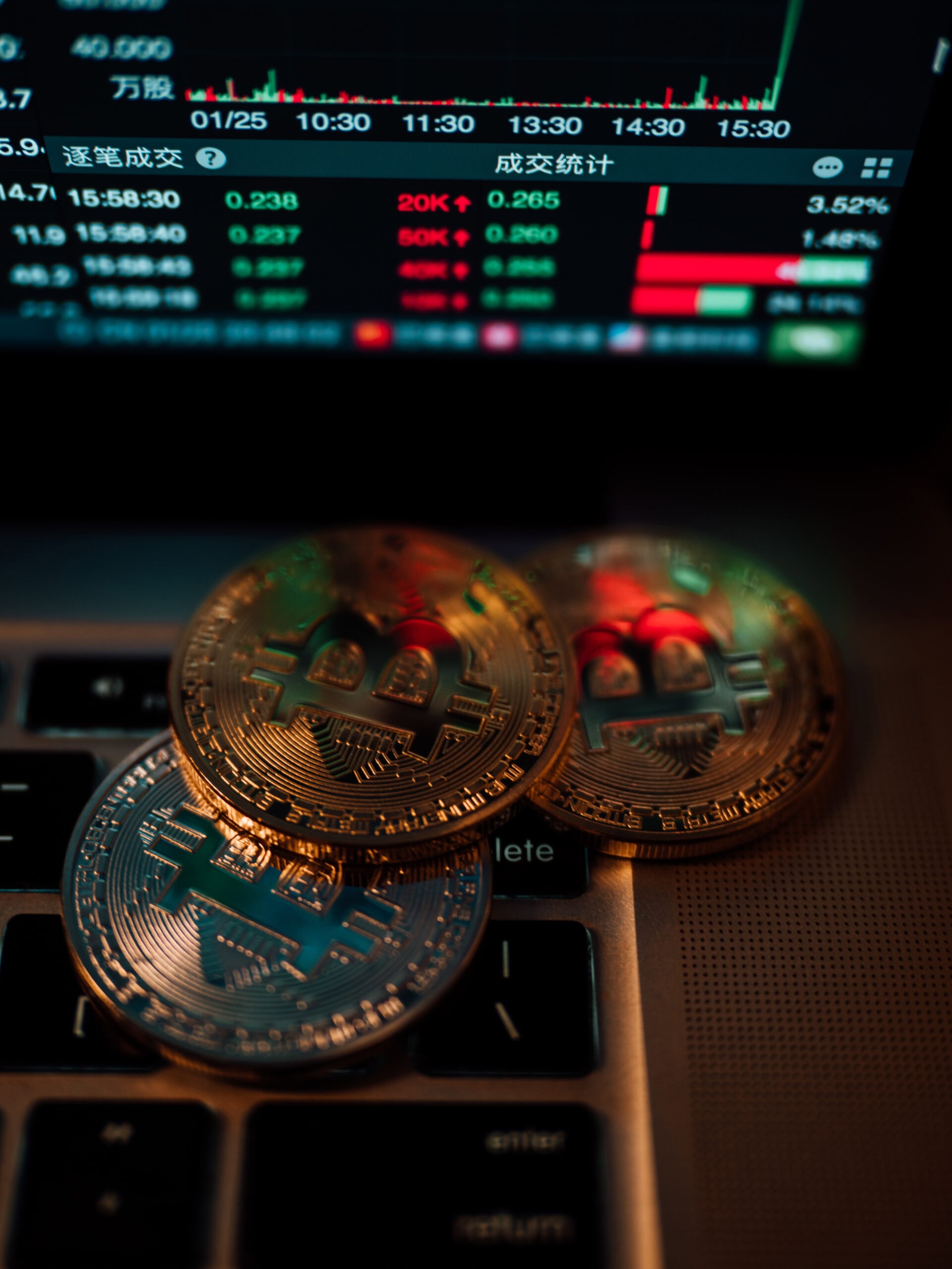 Three physical Bitcoin coins rest on a laptop keyboard, with a screen in the background displaying cryptocurrency trading data and charts in Chinese.