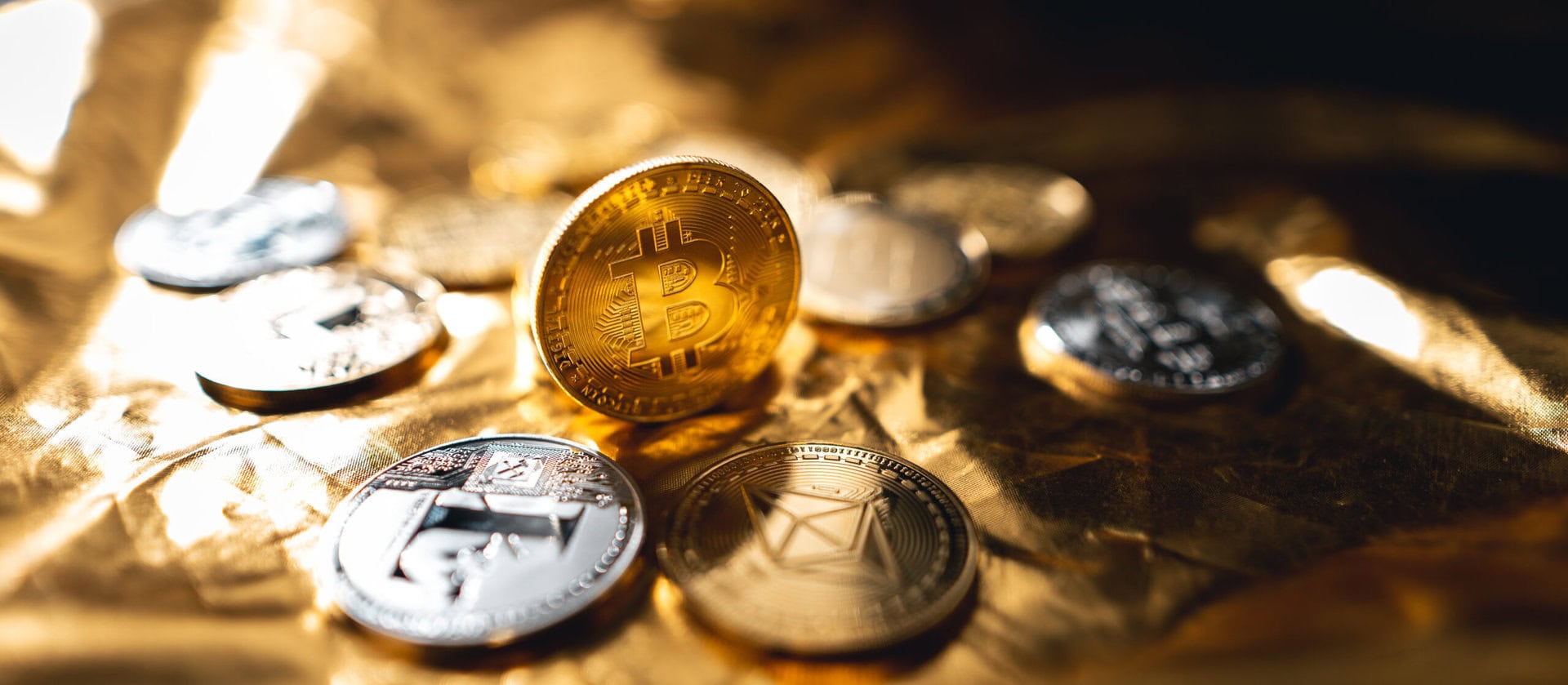 Several shiny gold and silver cryptocurrency coins, including a prominent Bitcoin coin standing upright, are displayed on a reflective gold surface.