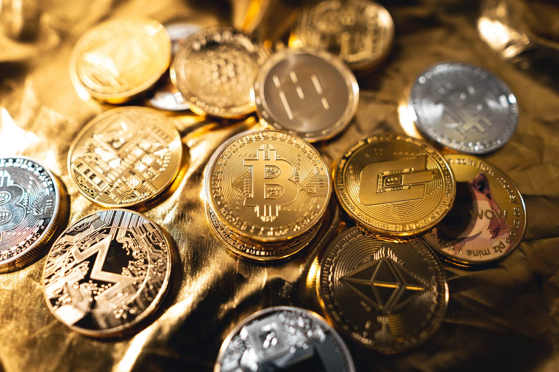 A close-up of various shiny gold and silver cryptocurrency coins, including Bitcoin, Ethereum, and Litecoin, displayed on a crumpled gold surface.