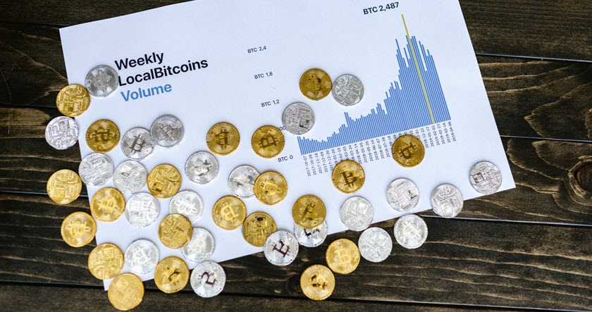 A printed chart showing weekly LocalBitcoins volume with scattered gold and silver bitcoin tokens on a wooden surface.