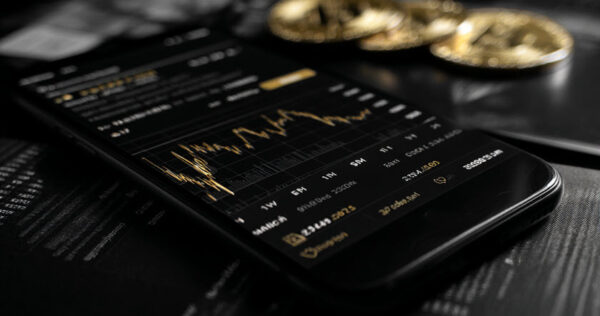 A smartphone displays a cryptocurrency price chart in a trading app, with physical coins resembling Bitcoin placed in the background.