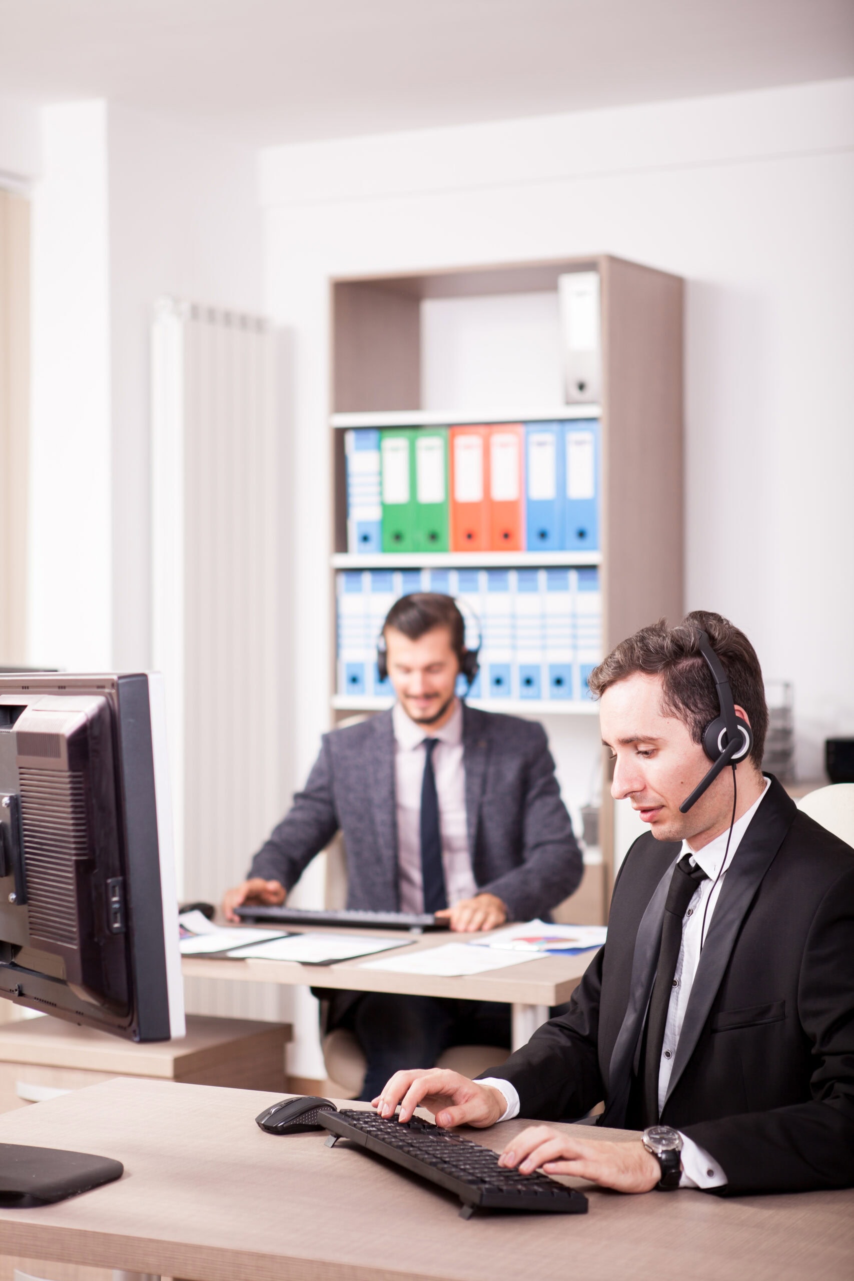 Two men in business attire sit at desks in an office, both wearing headsets and working on computers. Shelves with colorful binders are visible in the background.